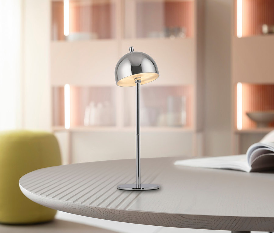 Silberfarbene SCHÖNER WOHNEN Akku-Tischleuchte »Bellis« mit kuppelförmigem Schirm auf einem hellen Tisch, im Hintergrund ein Magazin und ein gelber Pouf.