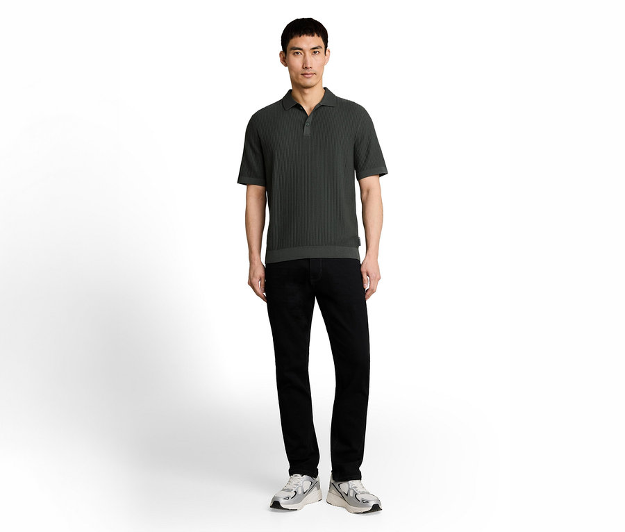 Ein Mann steht frontal vor weißem Hintergrund, trägt ein dunkelgrünes, geripptes Poloshirt, schwarze Jeans und grau-weiße Sneaker.