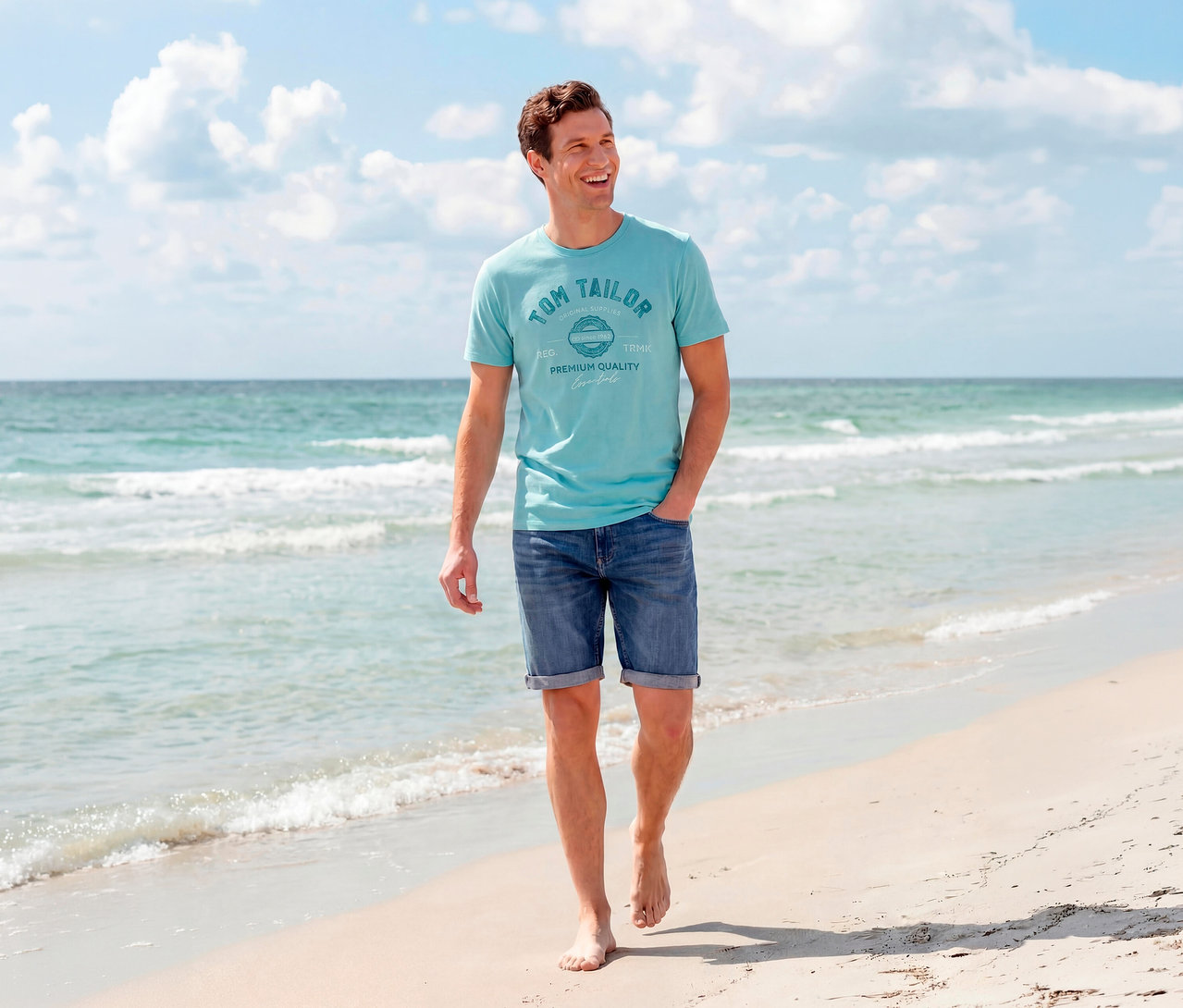 Ein lächelnder Mann geht barfuß am Sandstrand, trägt ein türkises Tom Tailor Logo T-Shirt und Tom Tailor TTJOSH REGULAR SLIM Jeans Shorts in Mid Stone Bright Blue Denim, mit Meer und Himmel im Hintergrund.