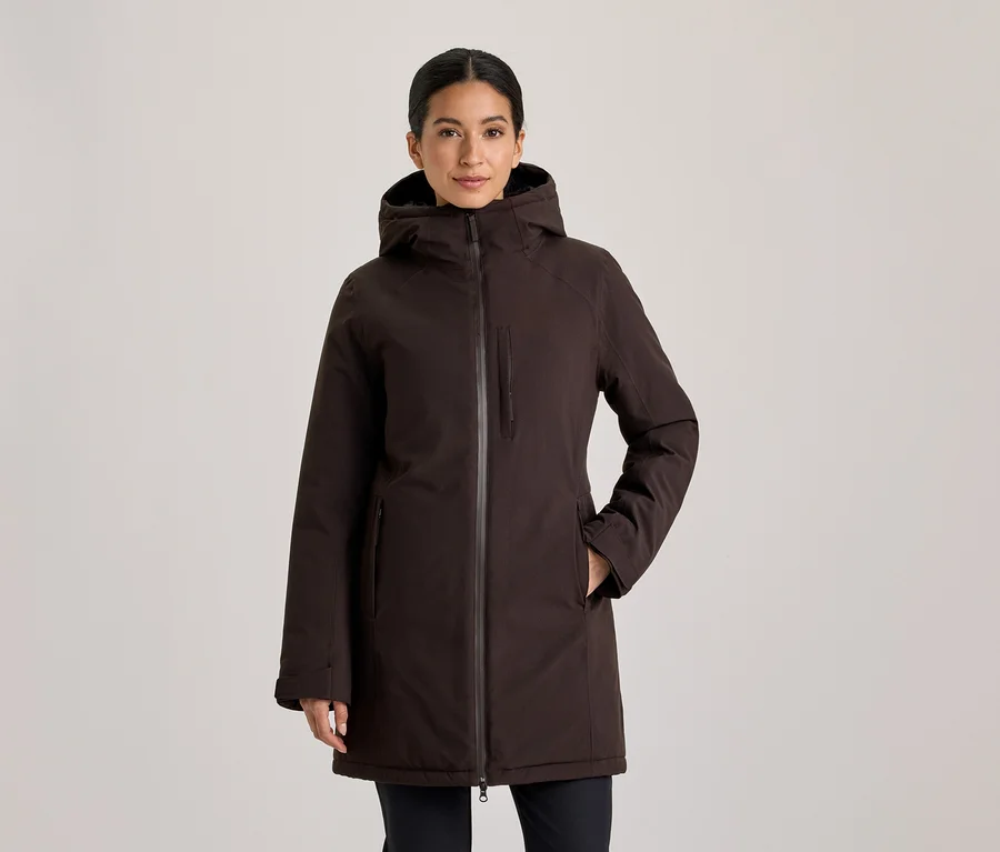 Frau mit zurückgebundenem dunklem Haar, die nach vorne blickt und eine dunkelbraune Kapuzenjacke mit den Händen in den Taschen trägt.