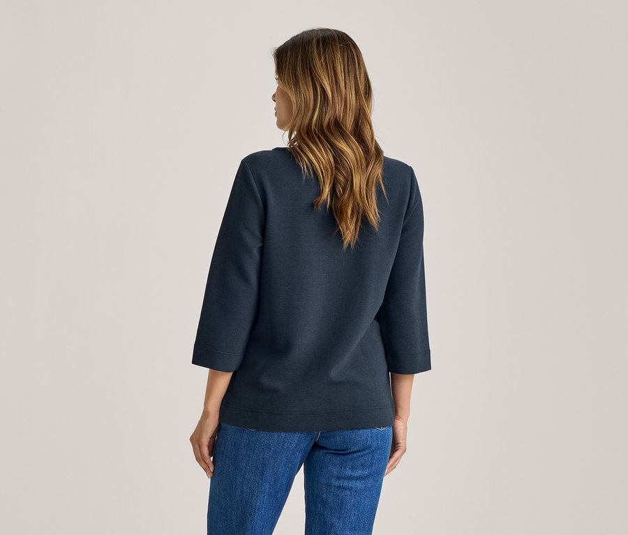 Frau von hinten, die ein dunkelblaues Oberteil mit Dreiviertelärmeln und blaue Jeans trägt. Sie hat langes, gewelltes braunes Haar.