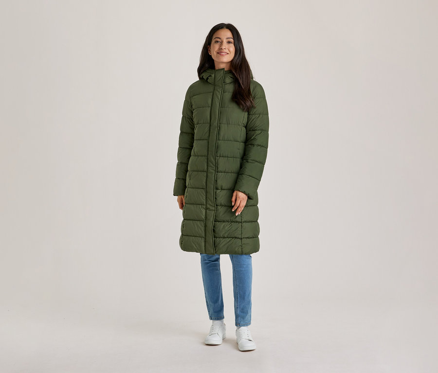 Eine lächelnde Frau mit dunklen Haaren steht frontal da und trägt einen langen, olivgrünen Steppmantel mit Kapuze, blaue Jeans und weiße Turnschuhe.