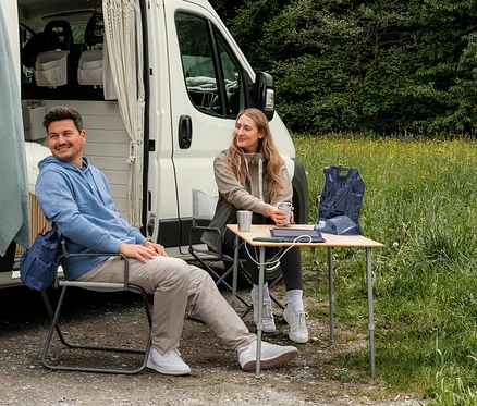 Ein Mann in einem hellblauen Sweat-Hoodie und eine Frau in einer beigen Sweatjacke und einer khakifarbenen Hybrid-Weste sitzen vor einem Campervan. Auf einem Tisch liegt eine dunkelblaue Pack-Weste.