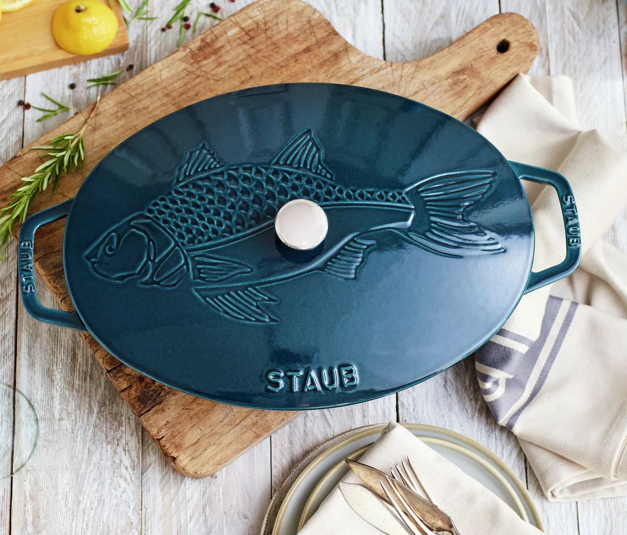Ein ovaler, petrolfarbener Staub Bräter mit erhabenem Fischdesign auf dem Deckel steht auf einem Holzschneidebrett. Daneben liegen gestapelte Teller mit Besteck und einer Serviette. Zitronen, Rosmarin und Pfefferkörner sind auf dem hellen Holztisch verteilt.