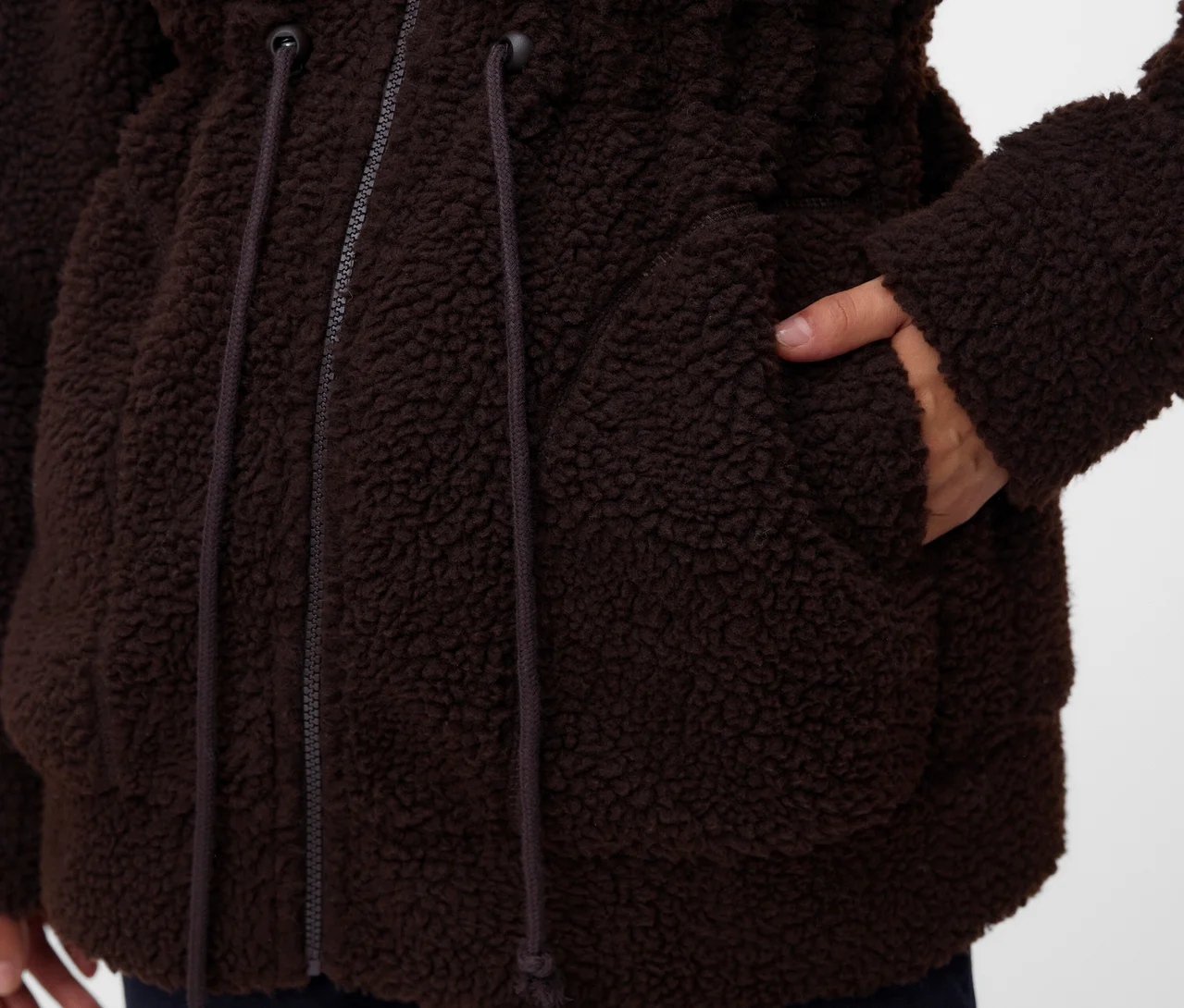 Nahaufnahme einer Person in einer braunen, flauschigen Jacke mit der Hand in einer Tasche. Die Jacke hat einen Reißverschluss und Kordelzüge an der Taille.