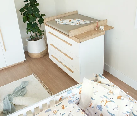 Ein Babyzimmer mit einer weißen Kommode und einem Wickeltischaufsatz aus Holz. Darauf liegen eine graue Wickelauflage und eine Decke mit Tiermotiven, daneben eine Giraffen- und Eisbärenfigur. Im Vordergrund steht ein weißes Babybett mit passender Bettwäsche. Eine große Pflanze in einem geflochtenen Korb steht neben der Kommode, ein heller Teppich liegt auf dem Holzboden.