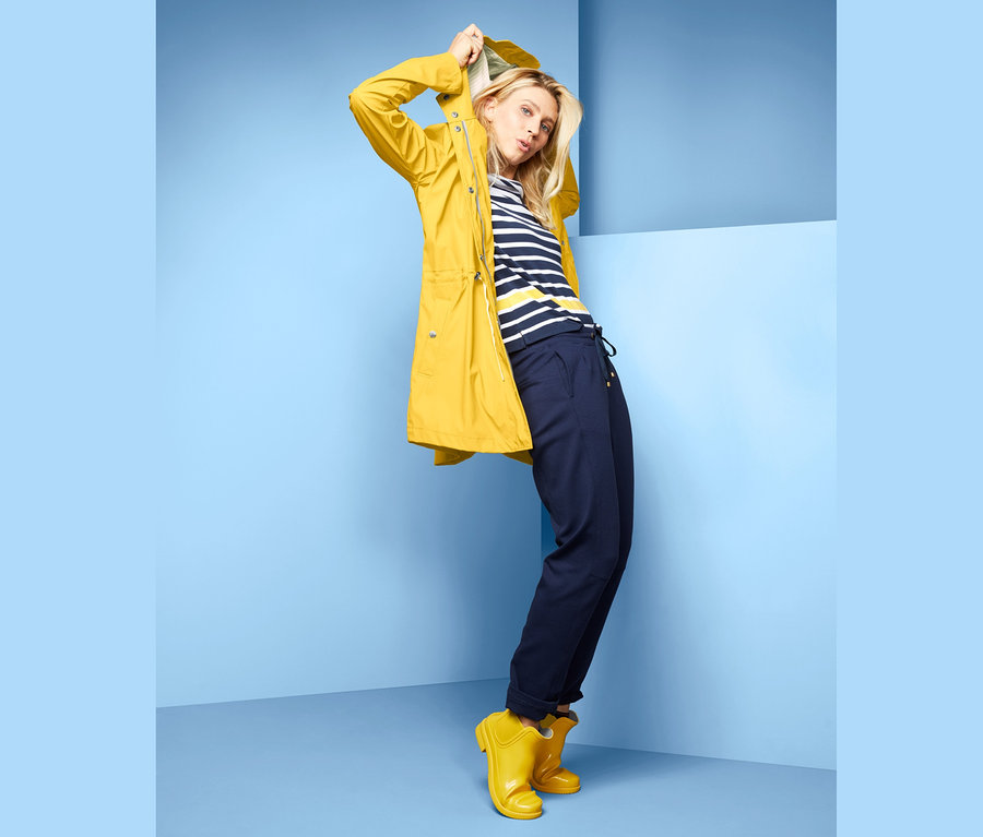 Frau posiert in gelbem Regen-Parka, gestreiftem T-Shirt und marineblauer Hose.