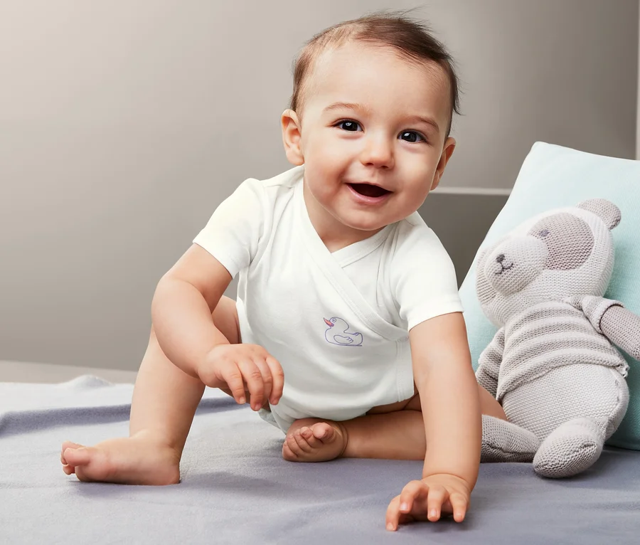 Lächelndes Baby sitzt auf einer Decke und trägt einen weißen Body. Neben ihm liegt ein Stofftier-Bär.