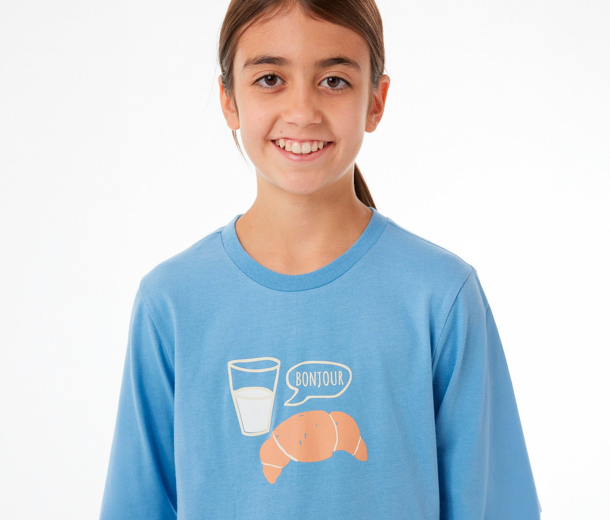 Lächelndes junges Mädchen in einem blauen SCHIESSER Kinder-Pyjama-Oberteil mit einem Aufdruck aus Milchglas, Croissant und einer Sprechblase mit „BONJOUR“.