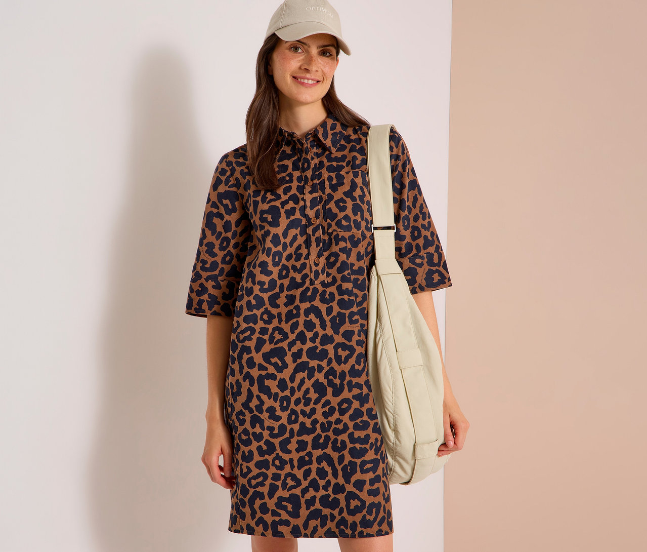 Lächelnde Frau in braun-schwarzem Leopardenkleid, beiger Baseballkappe und beiger Umhängetasche.