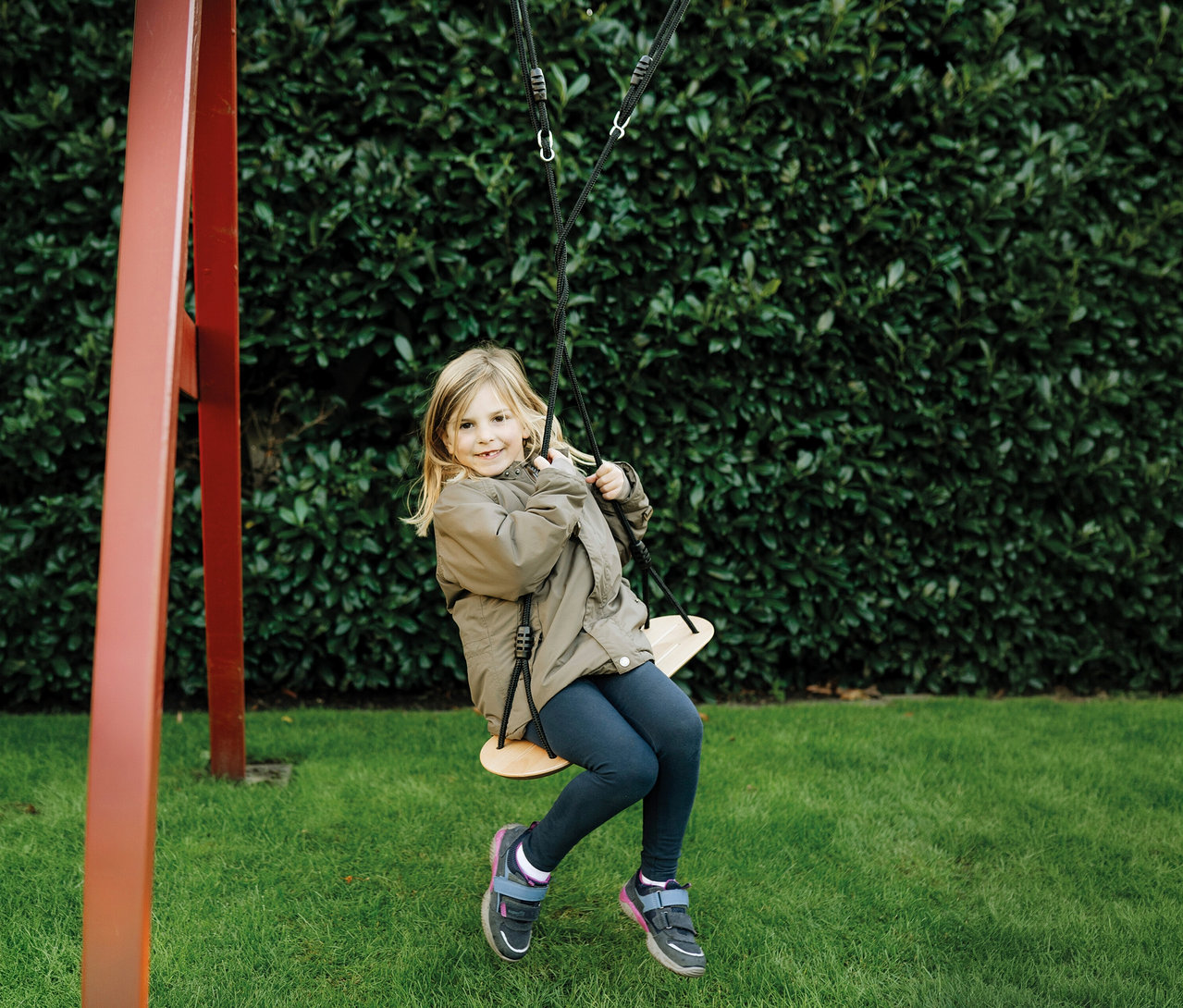 Ein junges Mädchen mit blonden Haaren lächelt, während es auf einer small foot XL-Brettschaukel sitzt, trägt eine braune Jacke und dunkle Leggings, mit einer grünen Hecke und Gras im Hintergrund.