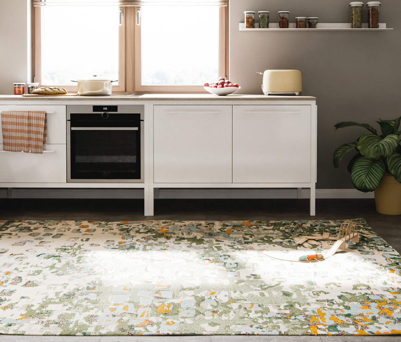 Eine moderne Küche mit weißen Schränken, heller Arbeitsplatte und einem großen Fenster. Auf der Arbeitsplatte stehen ein Topf, Brot, Obst und ein gelber Toaster. Ein Backofen ist in die Schränke integriert. Rechts befinden sich eine grüne Pflanze und ein Regal mit Gewürzgläsern. Ein benuta Teppich »Casa Multicolor« mit abstraktem grünem, gelbem und blauem Muster bedeckt den Boden, darauf ein Holzspielzeugeisenbahn-Set.