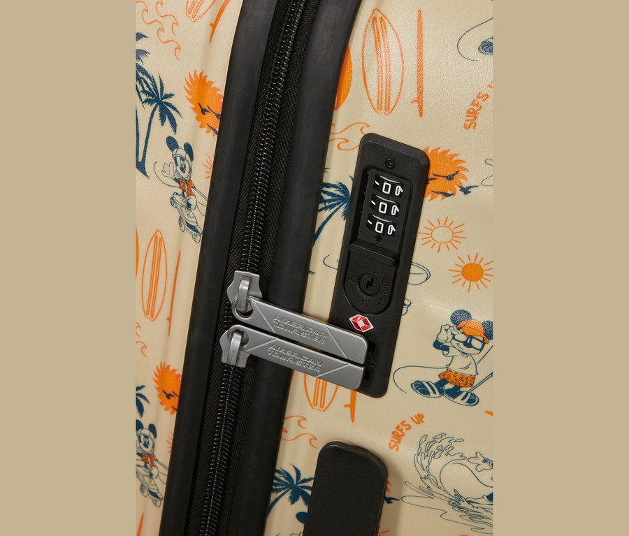 Nahaufnahme eines beigen Koffers mit einem Muster aus surfendem Mickey Maus, Palmen, Surfbrettern und Sonnen. Ein schwarzer Reißverschluss und zwei silberne "AMERICAN TOURISTER"-Reißverschlussgriffe sind an einem schwarzen Zahlenschloss mit TSA-Logo befestigt.