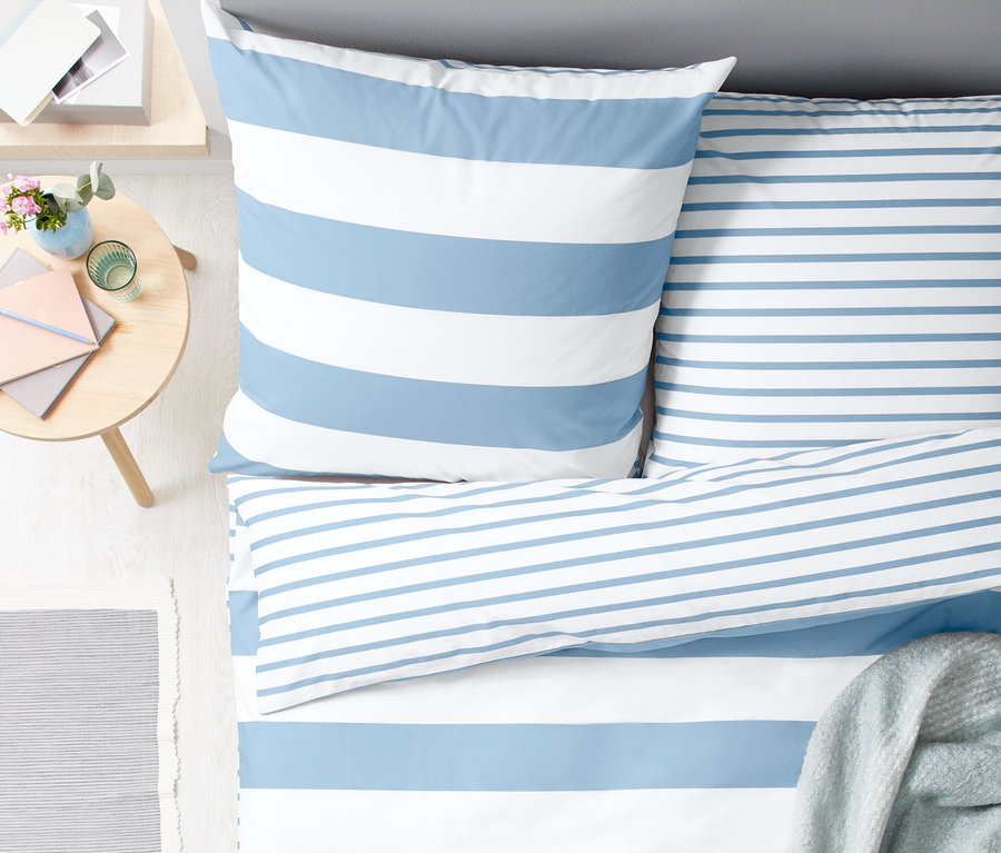 Draufsicht auf ein Doppelbett mit Perkal-Wendebettwäsche mit blauen und weißen Streifen. Eine graue Decke liegt auf dem Bett.