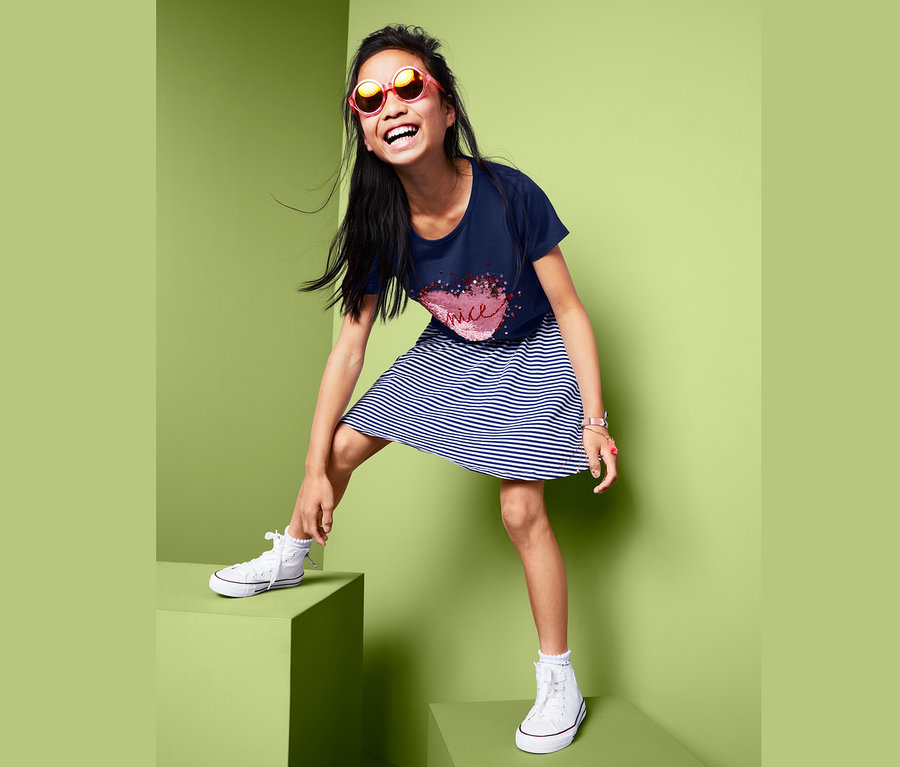 Ein lächelndes Mädchen in einem T-Shirt mit Wendepailletten, einem gestreiften Kleid und Turnschuhen steht auf grünen Würfeln.