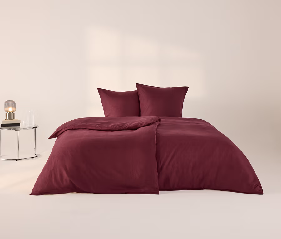 Ein Bett mit beerenfarbener Leinen-Bettwäsche, zwei Kissen und einer gefalteten Decke. Links steht ein runder Beistelltisch mit Lampe und Flasche.