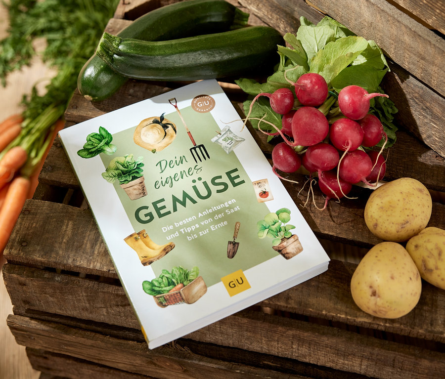 Das Buch »Dein eigenes Gemüse« liegt auf einer Holzkiste, umgeben von Karotten, Zucchini, Radieschen und Kartoffeln.