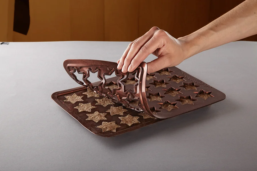 Eine Hand entfernt eine braune Silikon-Backform in Sternform von einem Blech mit sternförmigen Keksen.