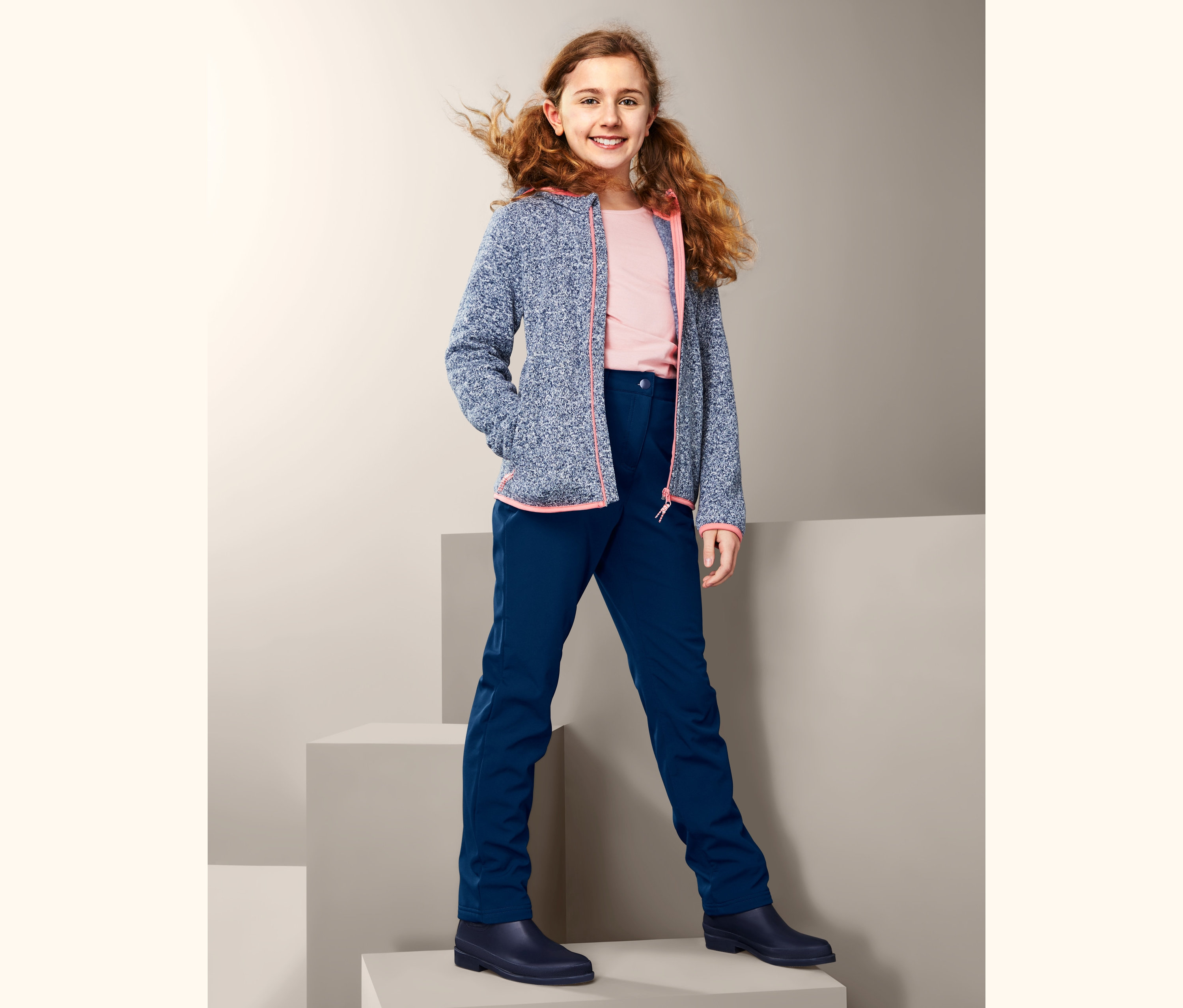 Lächelndes Mädchen posiert auf grauen Würfeln in blauer Softshellhose und blauer Jacke.