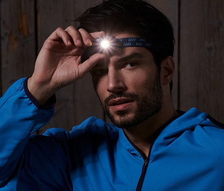 Mann sitzt auf einer Holzbank und justiert eine blaue, kompakte Stirnlampe.