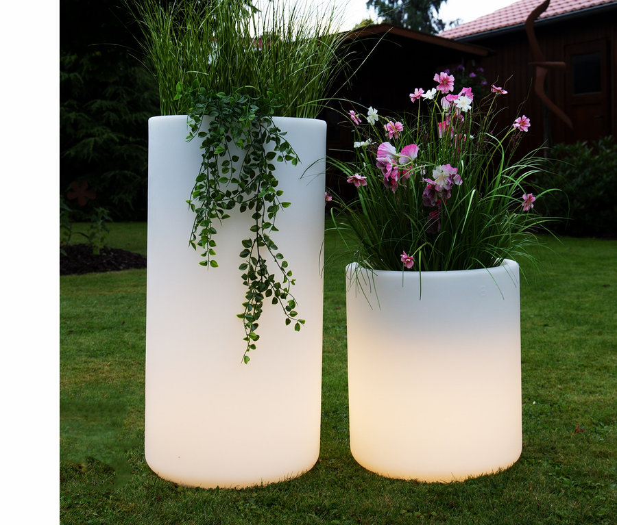 Zwei weiße Blumentöpfe stehen auf einer Wiese, einer mit Gras und Kletterpflanzen, der andere mit rosa und weißen Blumen. Im Hintergrund ist eine 8 seasons design LED-Leuchte zu sehen.