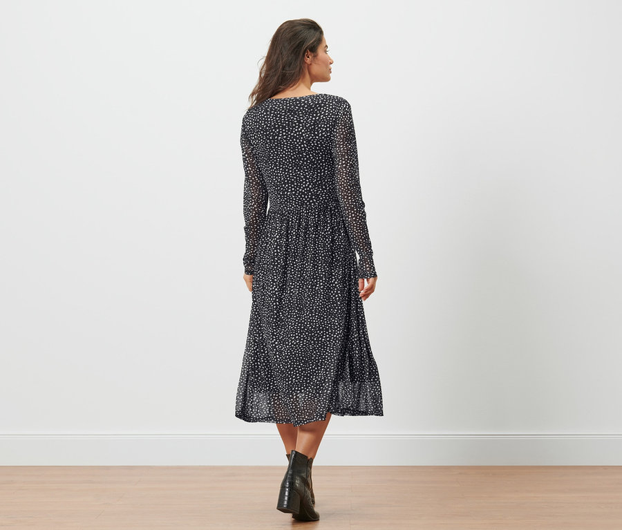 Eine Frau geht von hinten in einem gepunkteten Mesh-Kleid und schwarzen Stiefeln über den Boden.