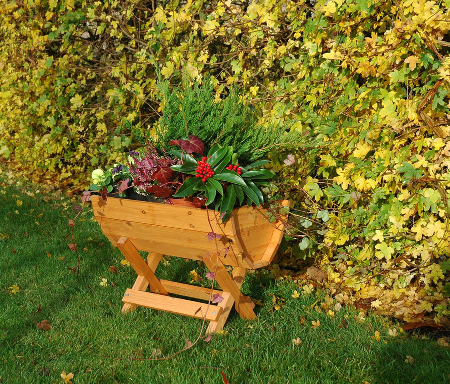 Ein Blumenfass aus Holz mit Pflanzen steht auf einem grünen Rasen vor einer Hecke.