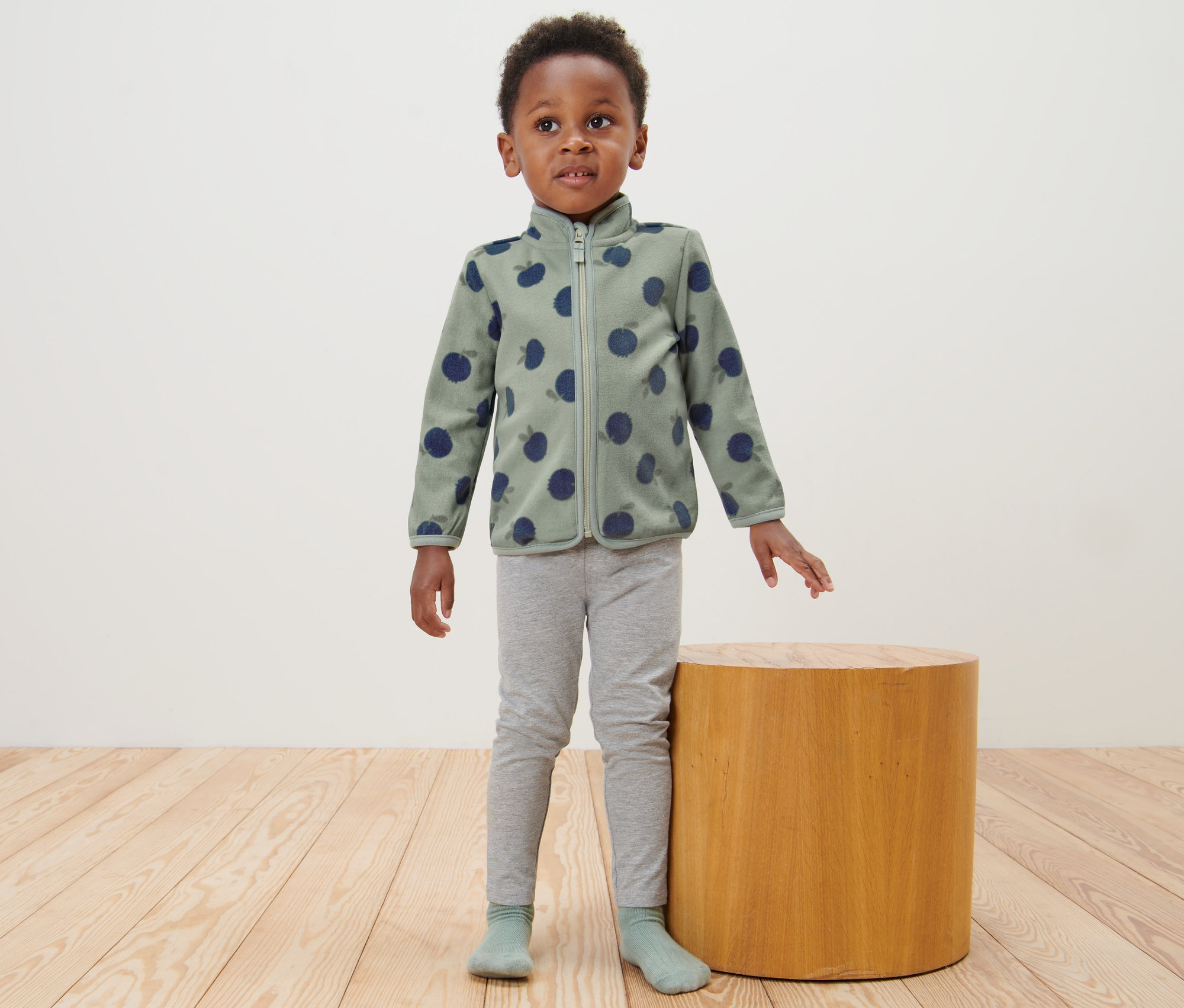 Ein Junge steht neben einem Holztisch und trägt eine Kinder-Fleecejacke, Blaubeere.