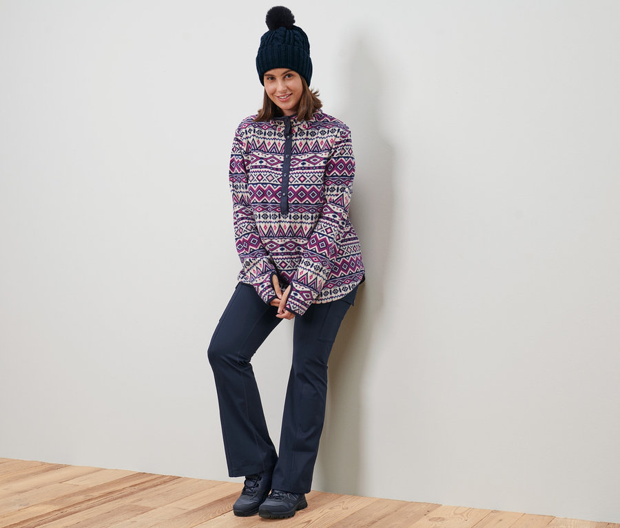 Eine Frau lehnt an einer Wand und trägt einen Fleecepullover, eine Strickmütze und eine Thermo-Jazzpants.