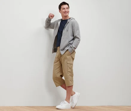 Mann lehnt an Wand, trägt beige Cargo-Bermuda in 3/4-Länge, T-Shirt, Kapuzenpullover und Turnschuhe.