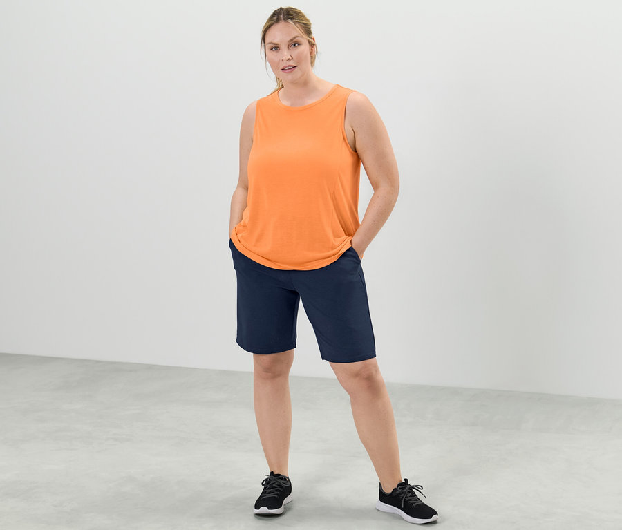 Eine Frau steht mit den Händen in den Hosentaschen, trägt ein pfirsichfarbenes Sport-Top aus einem zweiteiligen Set und dunkelblaue Shorts.