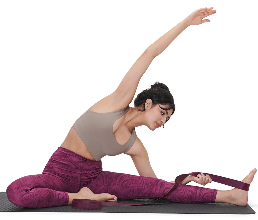 Frau sitzt auf einer Yogamatte und benutzt einen lila 2-in-1-Yogagurt, um ihr Bein zu dehnen.