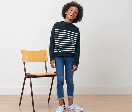 Ein lächelndes Kind steht neben einem Stuhl und trägt ein blaues Kinder-Sweatshirt.