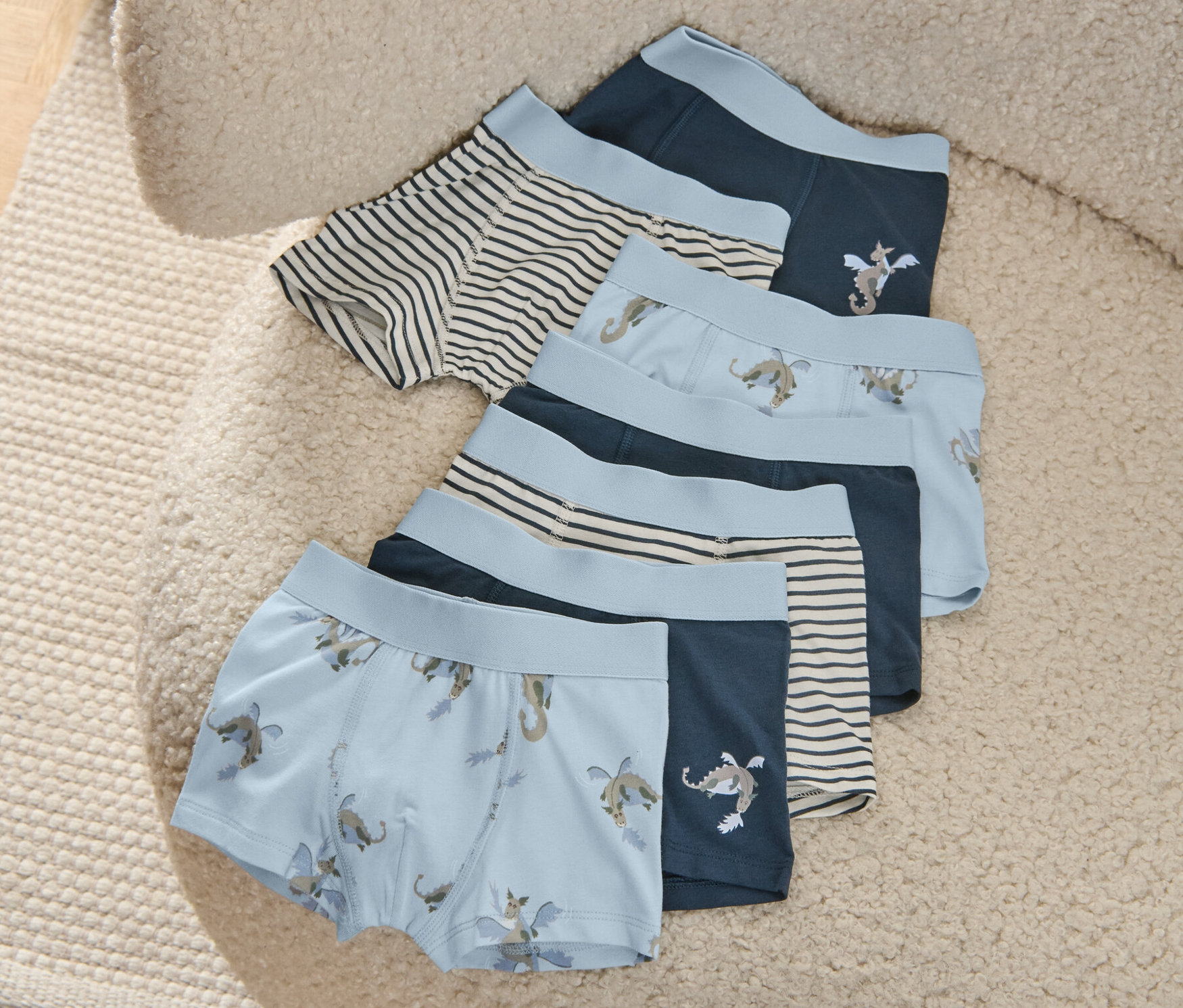 Ein 7er-Pack Kinder-Slipboxer liegt auf einem beigen, zotteligen Sessel. Die Boxershorts sind blau mit Drachenmotiven und Streifen.