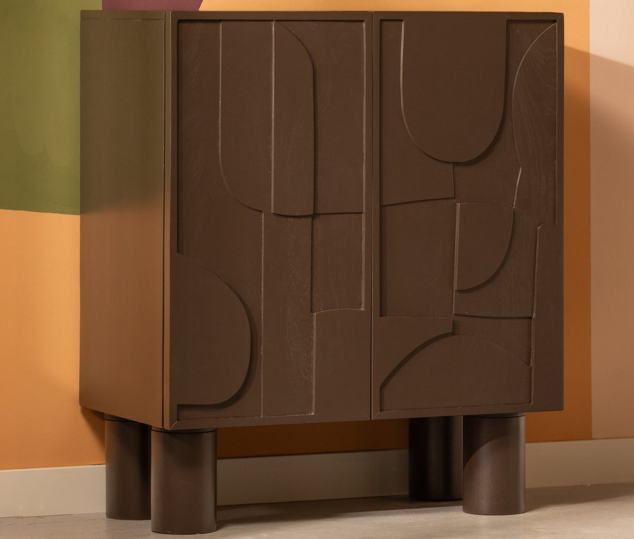 Ein brauner BePureHome Schrank »Notice« mit dekorativen geometrischen Formen auf den Türen steht auf dem Boden vor einer mehrfarbigen Wand.