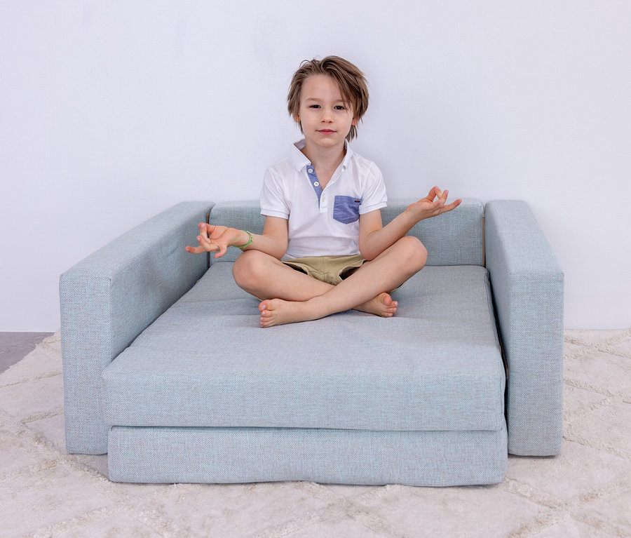Junge sitzt im Schneidersitz in Lotusposition auf mintgrauem Schlafsofa.