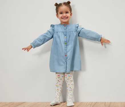 Ein lächelndes Mädchen im Jeanskleid steht mit ausgestreckten Armen und trägt eine 5 Kinder-Strumpfhosen, floral.