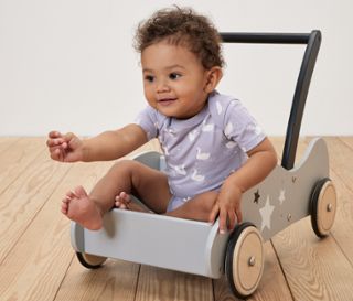 Lockiges Kleinkind sitzt in einer grauen Lauflernhilfe aus Holz mit Sternenmuster und trägt einen 5er-Pack Baby-Bodys mit Mitwachsfunktion, Prinzessin.