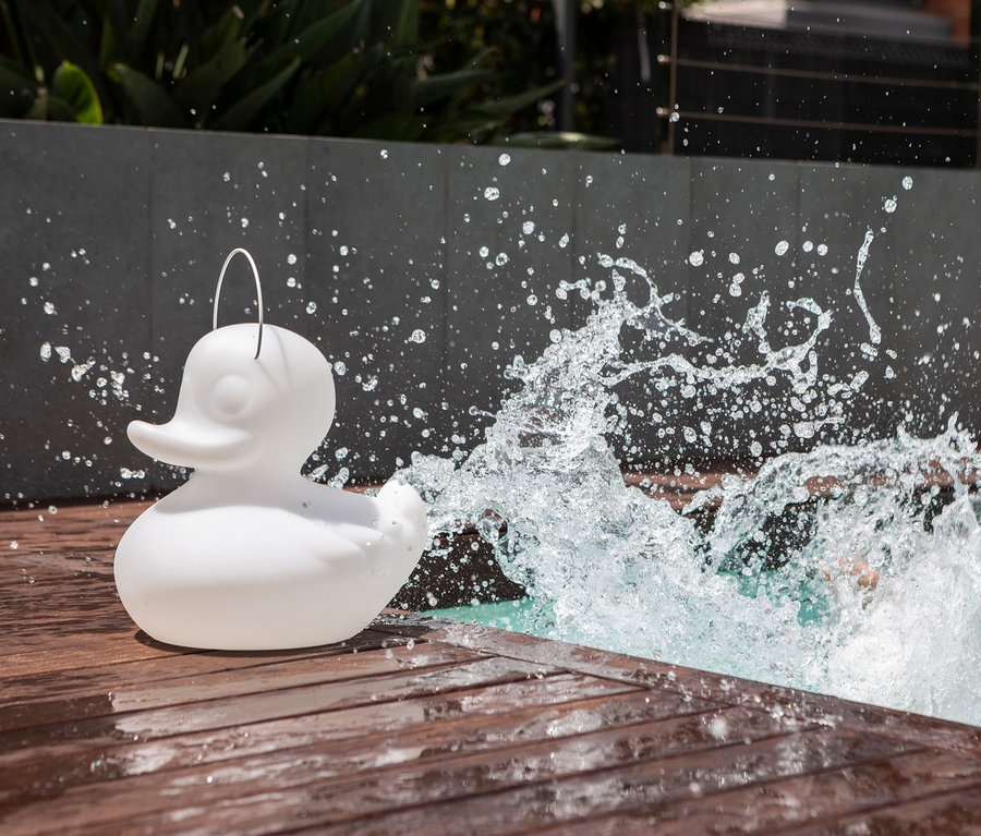 Auf einer Holzterrasse am Pool steht eine weiße Goodnight Light LED-Leuchte »Ente« S, die mit Wasser bespritzt wird.