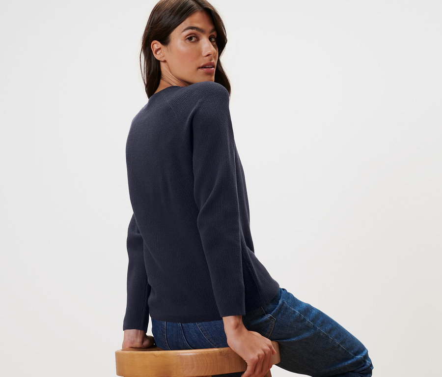 Frau sitzt seitlich auf einem Holzstuhl und blickt über die Schulter. Sie trägt einen dunkelblauen Strickpullover.