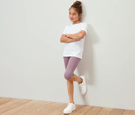 Mädchen in weißem T-Shirt und lila 3/4-Leggings (3 Kinder-3/4-Leggings) lehnt an der Wand.
