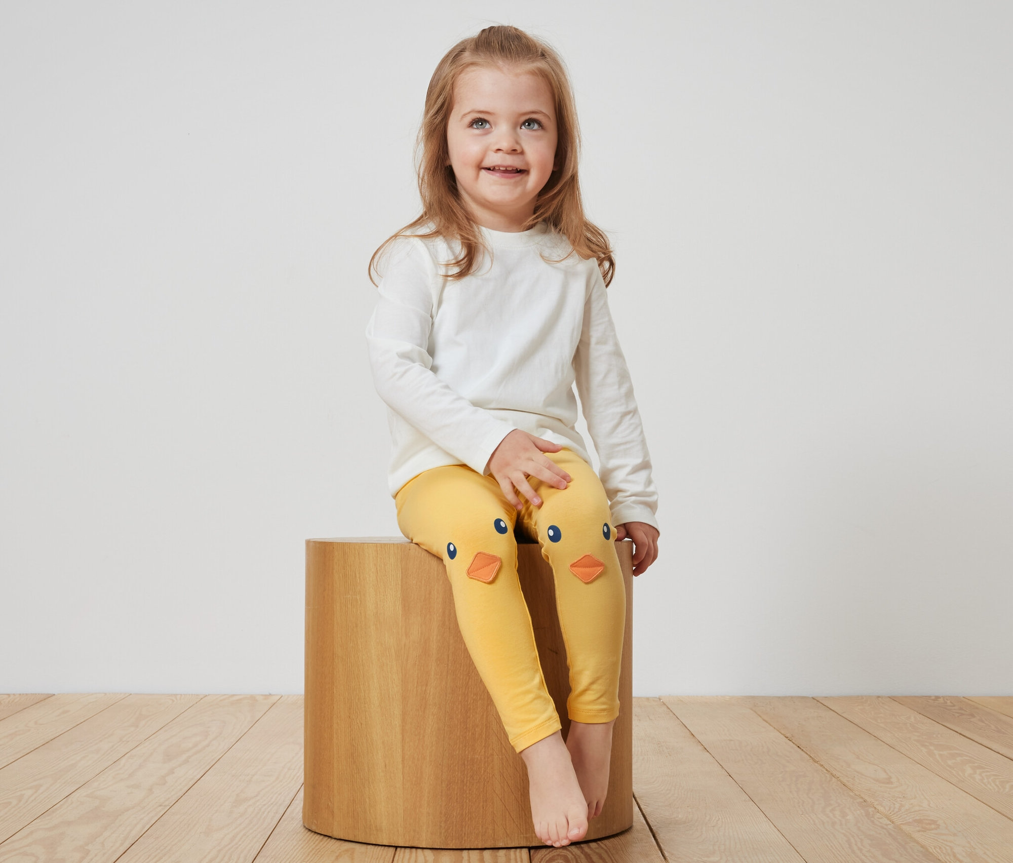 Ein Mädchen sitzt auf einem Holzhocker und trägt gelbe Kinder-Leggings mit Küken.