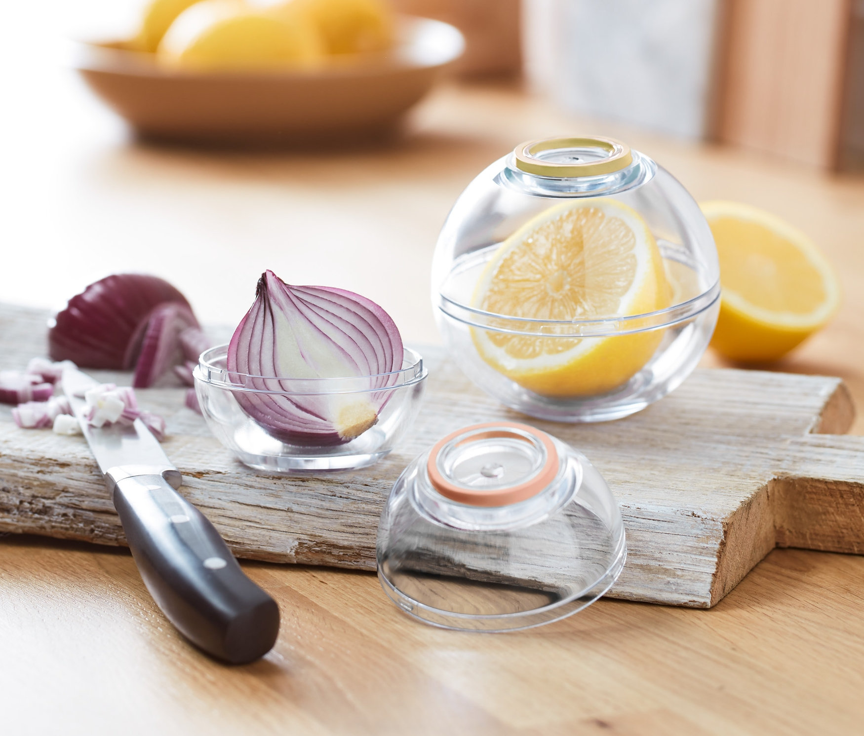 Eine halbe Zwiebel in einer runden Dose auf einem Holzbrett neben gehackten Zwiebeln, einem Messer und 2 Frischhaltekugeln mit Zitrone.