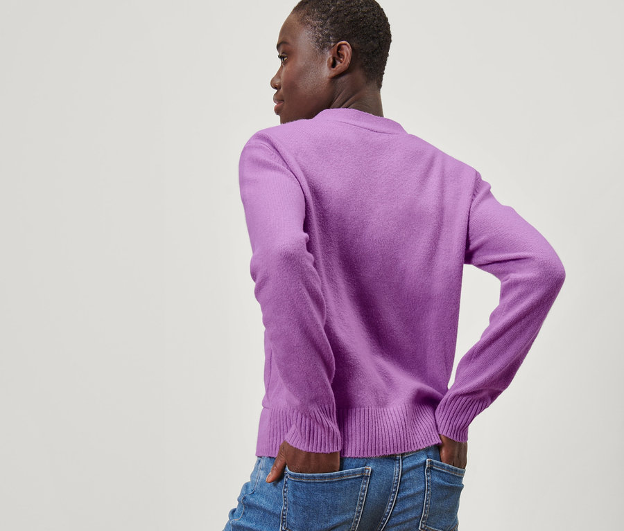 Schwarze Frau steht mit dem Rücken zur Kamera und hat die Hände in den Jeanshosentaschen. Sie trägt eine Strickjacke.