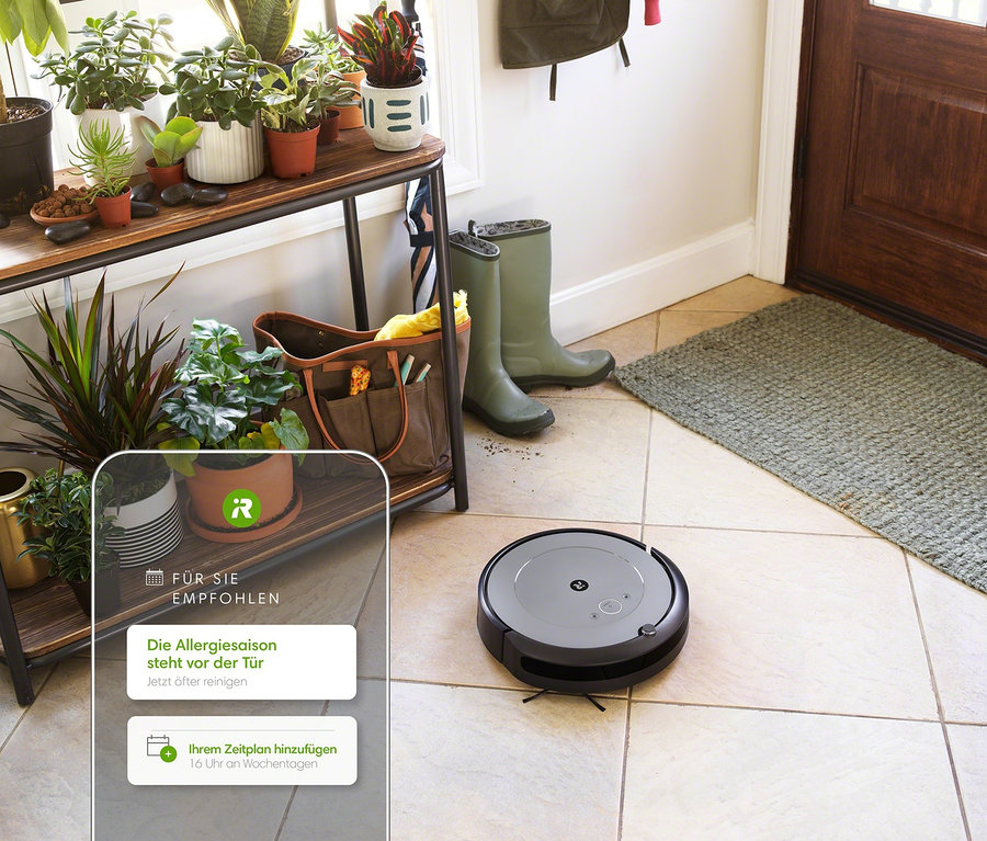 iRobot Saugroboter »Roomba i1« reinigt Fliesenboden im Flur. Im Hintergrund Regal mit Pflanzen und Stiefel.