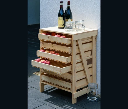 Ein Dobar Vorratsregal aus Holz mit drei Schubladen voller roter Äpfel, auf der Oberseite zwei Weinflaschen und zwei Gläser.