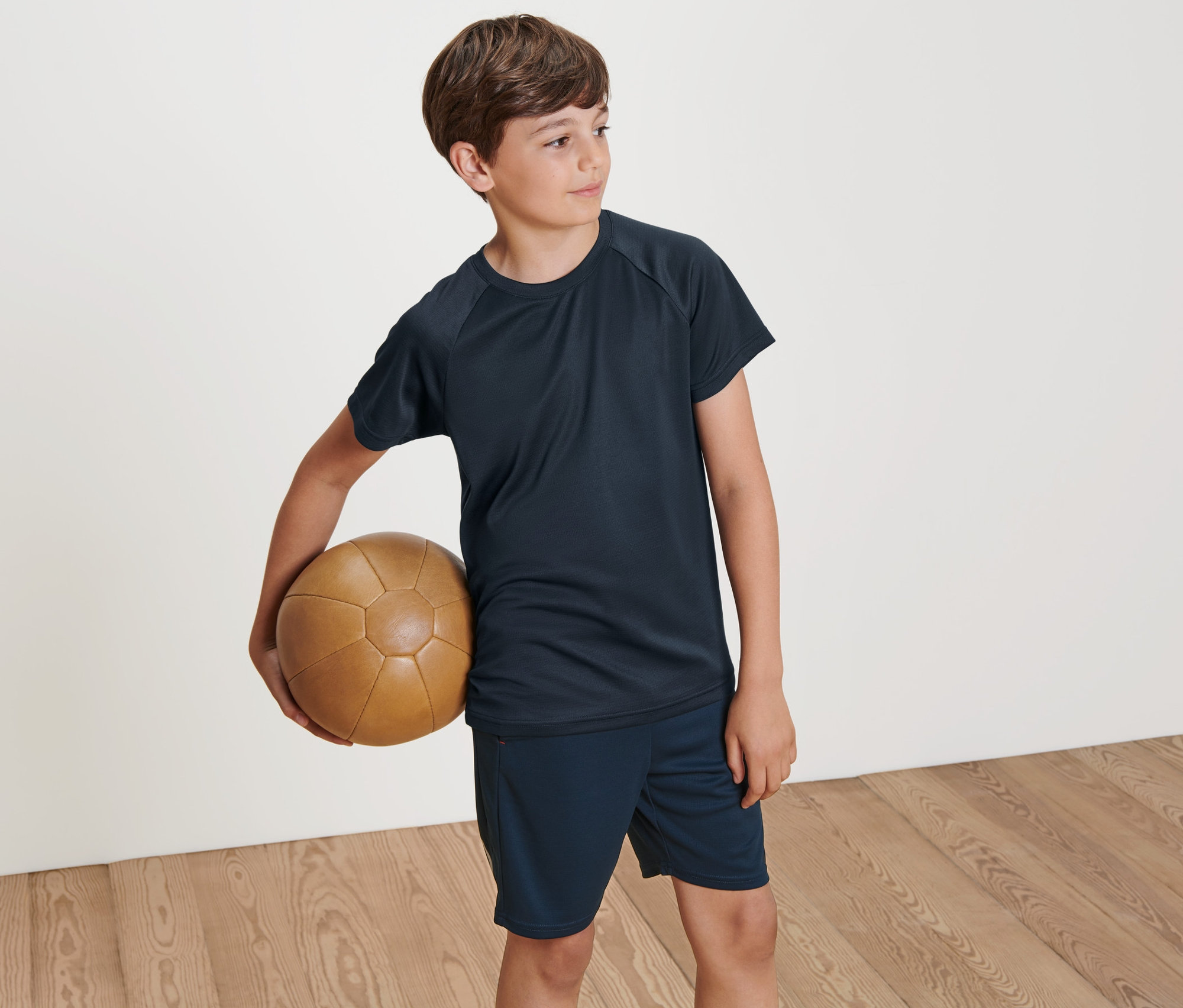 Ein Junge hält einen braunen Lederball. Er trägt eines von zwei Kinder-Funktionsshirts.