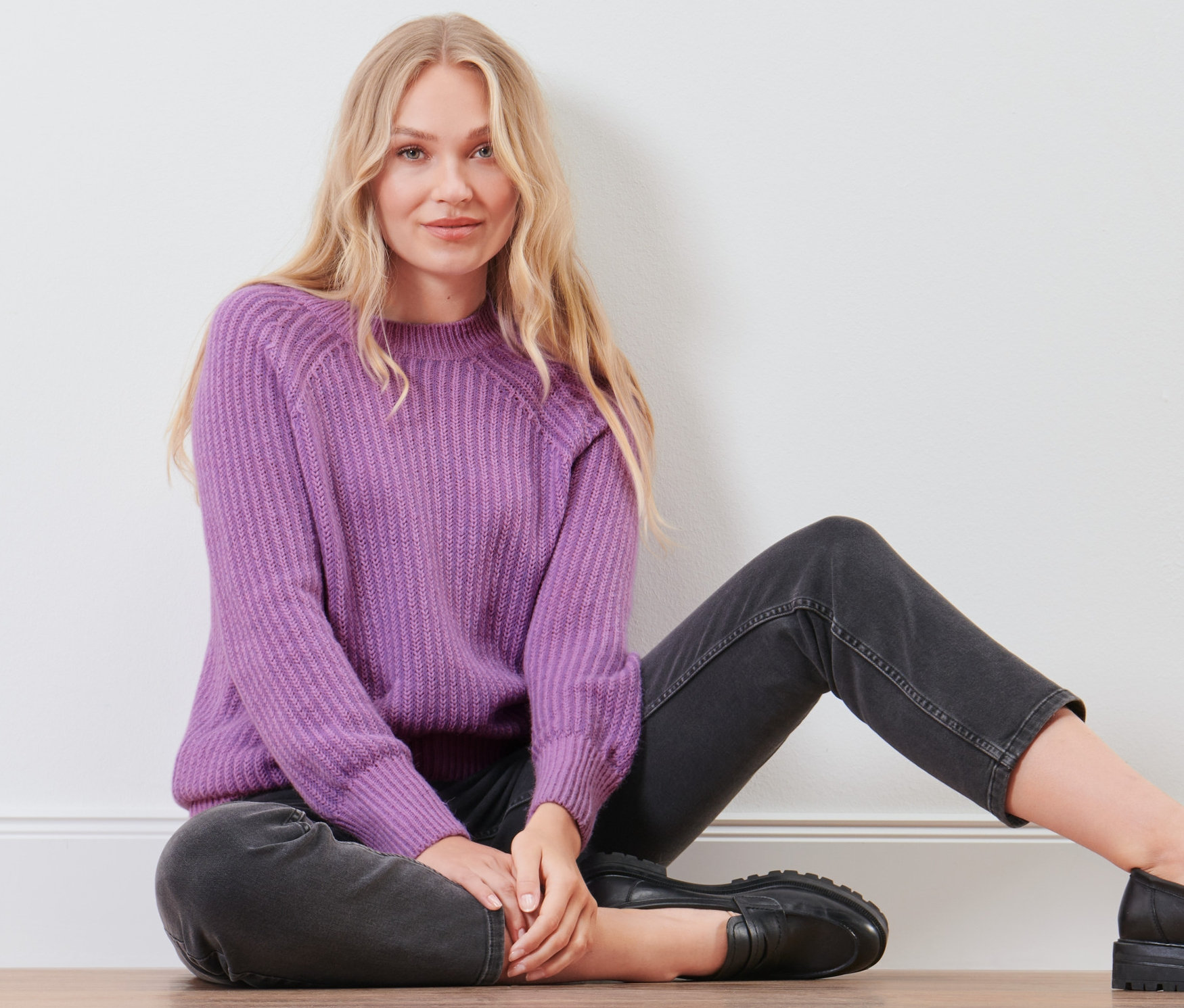 Eine blonde Frau sitzt auf dem Boden und trägt einen fliederfarbenen Strickpullover.