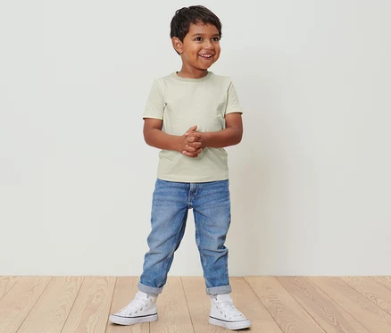 Ein Junge steht und trägt 2 Kinder-T-Shirts, Jeans und Turnschuhe.
