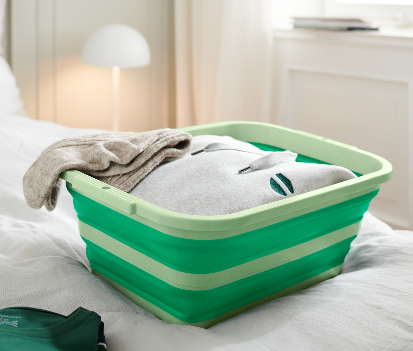 Ein grüner, faltbarer Wäsche-und-Spülkorb steht auf einem Bett. Der Korb ist mit Kleidung gefüllt.